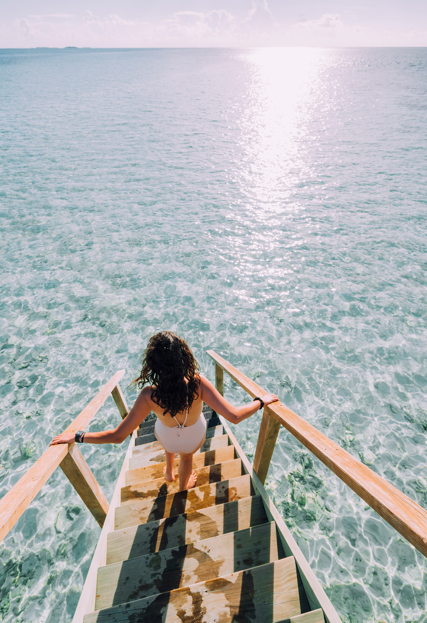 Woman walking down wooden stairs into crystal clear ocean water.